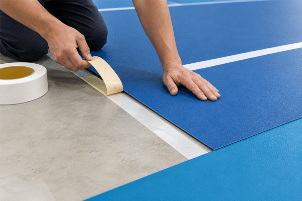 Worker installing blue pickleball court flooring with double sided tape