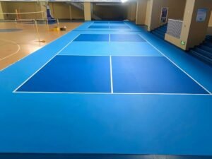 An interior photograph of an indoor sports facility, showing two newly installed blue and light blue roll-out pickleball courts side-by-side, with white boundary lines. In the background, there is a wooden-floored sports court area and tiered blue seating.